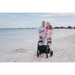 An elderly couple stands on a sandy beach by the sea. The man gestures and speaks, as the woman leisurely uses her Rollz Motion Performance All-Terrain 2-In-1 Rollator Transport Chair with air-filled tires. Both dressed for cool weather, they enjoy the seaside view.