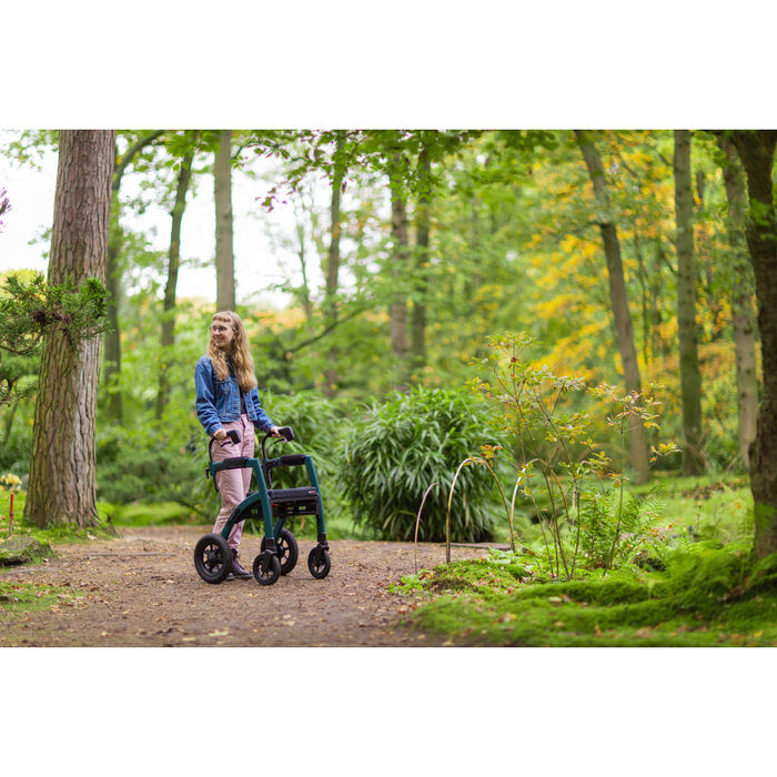 A woman with long hair navigates a forest path using the Rollz Motion Performance All-Terrain 2-In-1 Rollator Transport Chair. Dressed in a denim jacket and patterned pants, she enjoys a serene setting enhanced by air-filled tires for shock absorption among lush plants and trees.