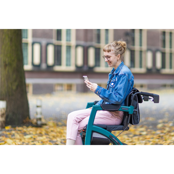 A person with glasses and light hair sits in a Rollz Motion Performance All-Terrain 2-In-1 Rollator Transport Chair with rear air-filled tires, using a smartphone. They wear a denim jacket, pink pants, and sit on autumn leaves with a building in the background.