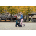 Wearing a denim jacket and pink pants, a person navigates effortlessly along a cobblestone street with the Rollz Motion Performance All-Terrain 2-In-1 Rollator Transport Chair. Autumn trees fill the scene, complemented by closed umbrellas over outdoor tables.