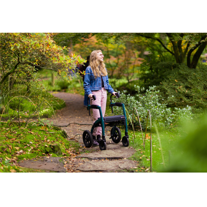 A woman with long hair, wearing a denim jacket and pink pants, uses the Rollz Motion Performance All-Terrain 2-In-1 Rollator Transport Chair with rear air-filled tires and shock absorption. She strolls through a lush garden path, gazing up amidst trees and shrubs.