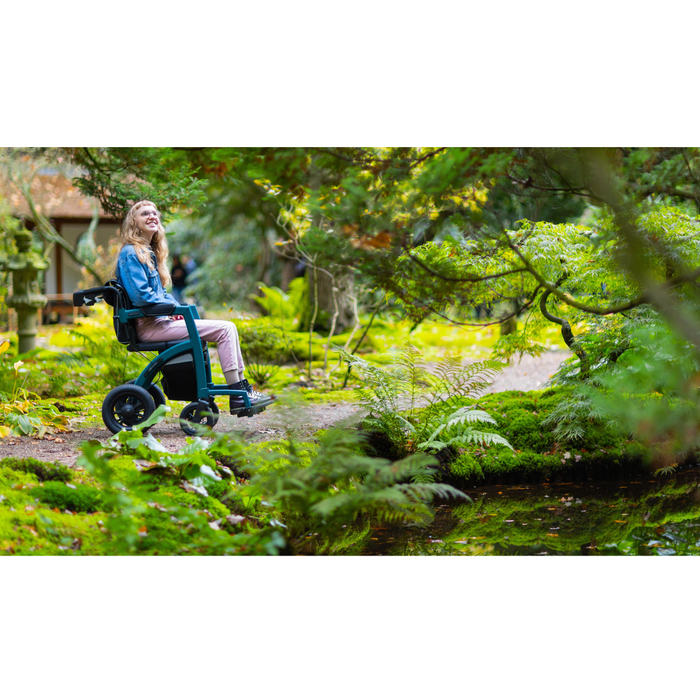 A person using the Rollz Motion Performance All-Terrain 2-In-1 Rollator Transport Chair smiles while exploring a lush garden. The air-filled tires and dirt pathway are surrounded by green ferns and trees, creating a serene atmosphere with excellent shock absorption for a smooth journey.