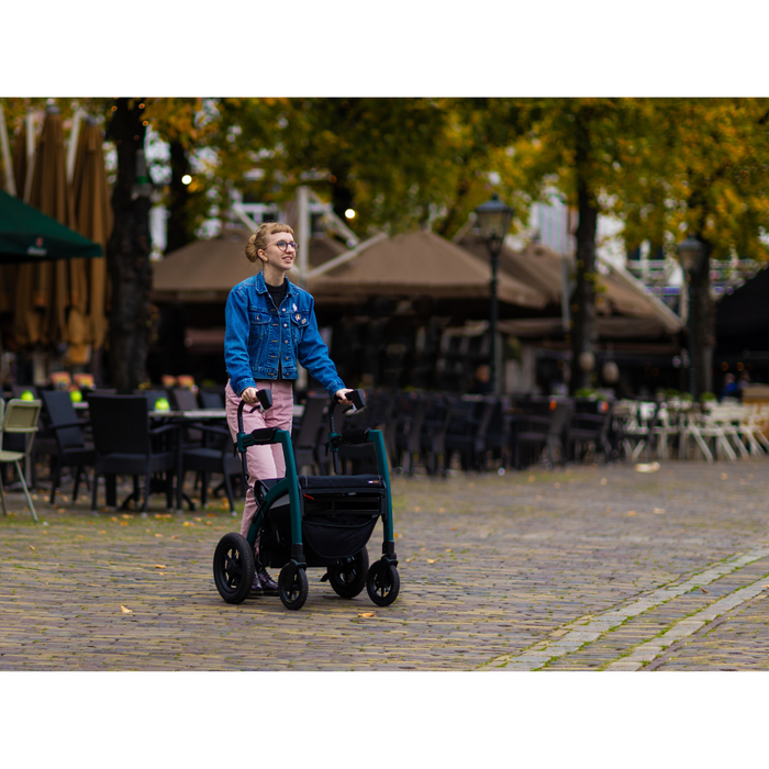 Sporting short hair, a blue denim jacket, and pink pants, a person expertly maneuvers the cobblestone street with the Rollz Motion Performance All-Terrain 2-In-1 Rollator Transport Chair. The backdrop showcases outdoor seating and trees decorated in vibrant fall colors.