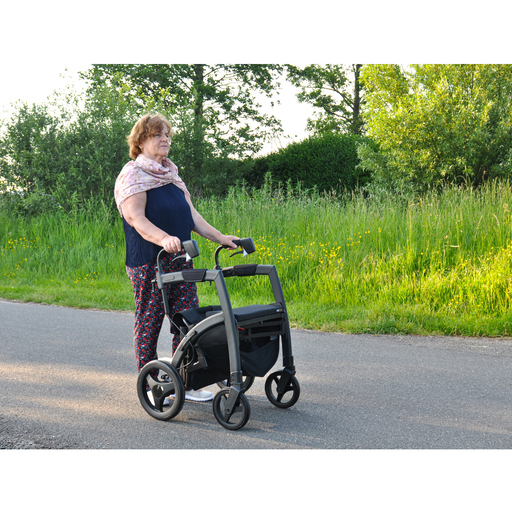 An elderly woman walks along a path using the Rollz Motion Rhythm 2-in-1 Parkinson Rollator Transport Chair with large wheels. Dressed in a dark top, floral pants, and pink scarf, she finds serenity amid lush greenery as she navigates life with neurological conditions.