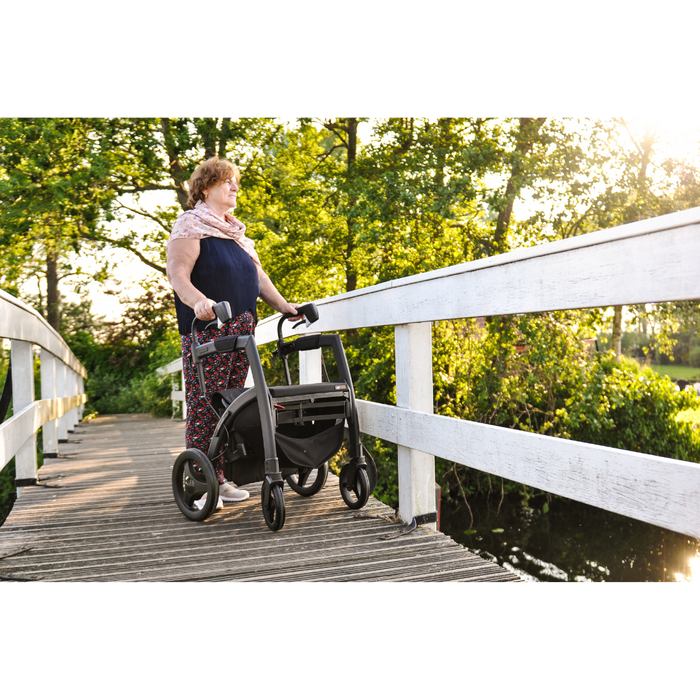 A woman stands on a wooden bridge in a sunlit park, using the Rollz Motion Rhythm 2-in-1 Parkinson Rollator Transport Chair with 1 Laser Cue. Shes surrounded by green trees and water below, appearing content as she enjoys the bright, tranquil setting despite neurological conditions.