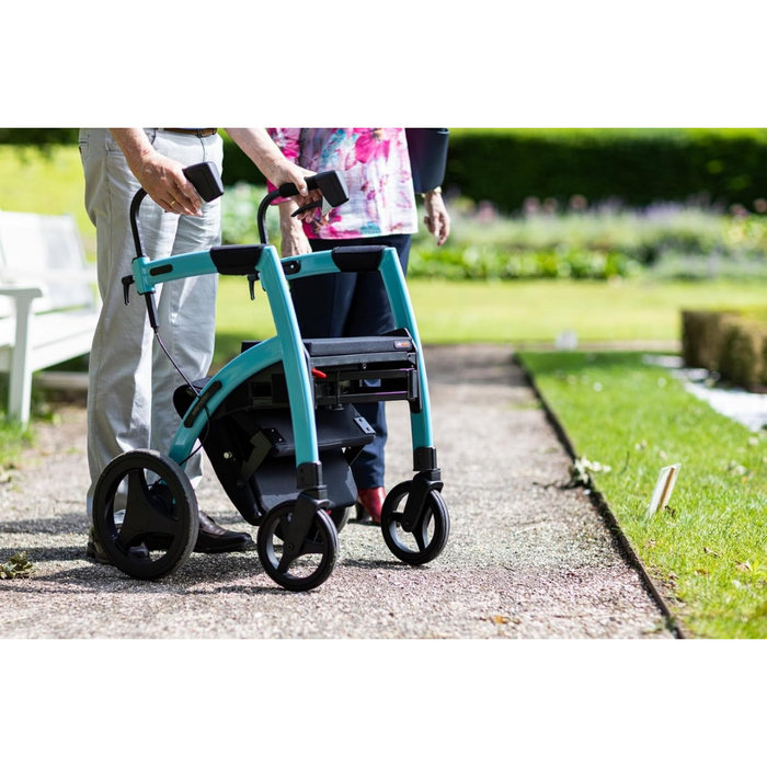 Two people, one with a teal Rollz Motion 2-in-1 Rollator Walker and Wheelchair, stroll on a sunny garden path beside green grass and blooming flowers.