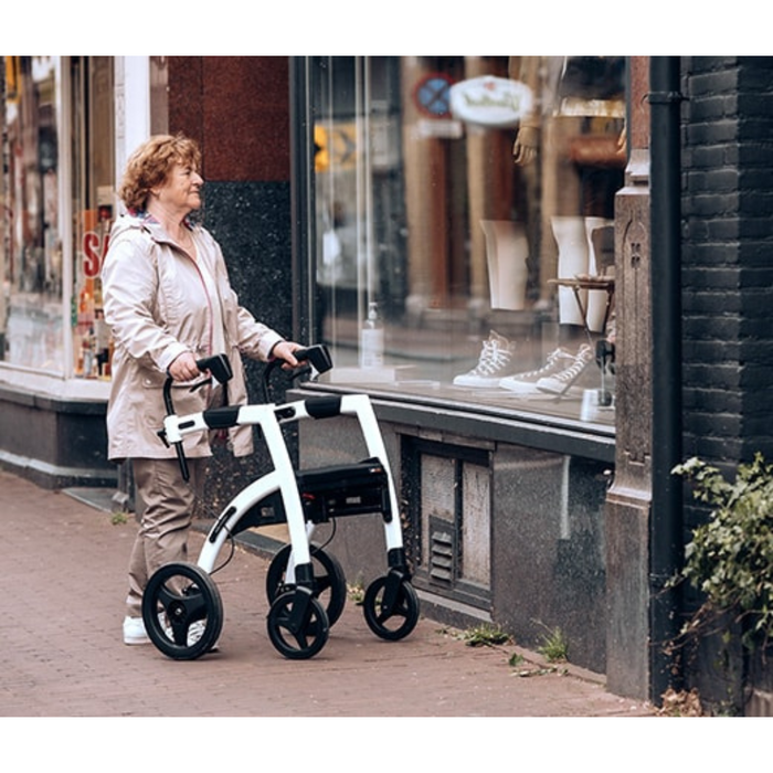 An elderly woman with short curly hair uses a Rollz Motion 2-in-1 Rollator Walker and Wheelchair while looking into a store window. Shes dressed in a light pink jacket and khaki pants, standing on a brick sidewalk in an urban area.