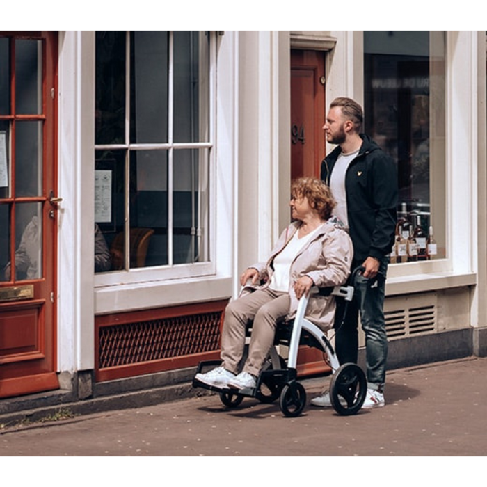 A man stands beside an older woman seated in a wheelchair on a sidewalk, in front of a building with large windows framed in red. He is pushing the Rollz Motion 2-in-1 Rollator Walker and Wheelchair, and both are gazing to the left.