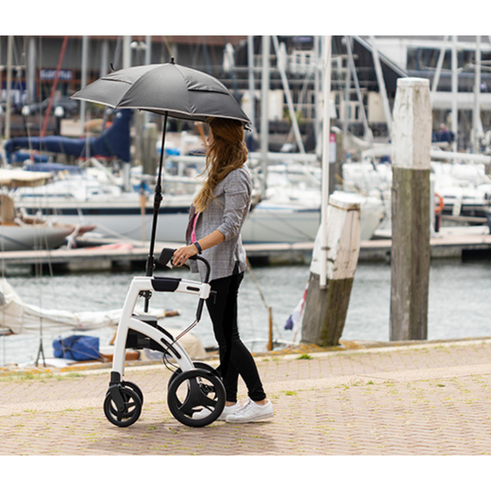 A person strolls along a dock with a Rollz Motion 2-in-1 Rollator Walker and Wheelchair, a white mobility aid with large wheels and an attached umbrella. Boats and masts grace the water in the background. The individual wears a gray top and black pants, perfectly blending comfort with functionality.