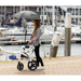 A person strolls along a dock with a Rollz Motion 2-in-1 Rollator Walker and Wheelchair, a white mobility aid with large wheels and an attached umbrella. Boats and masts grace the water in the background. The individual wears a gray top and black pants, perfectly blending comfort with functionality.