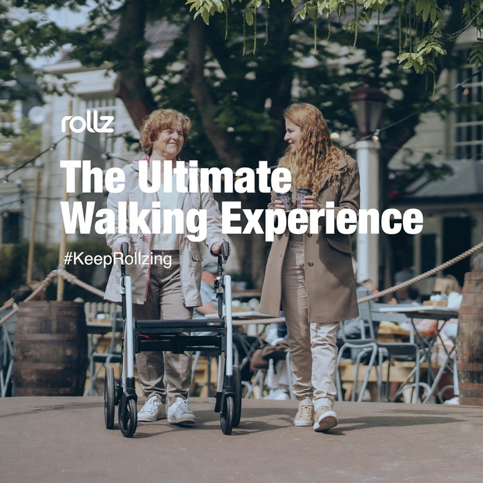 Two women walk along a tree-lined path by an outdoor cafe. One uses a Rollz Motion 2-in-1 Rollator Walker and Wheelchair, and they seem engaged in conversation. The text reads, The Ultimate Walking Experience and #KeepRollzing.