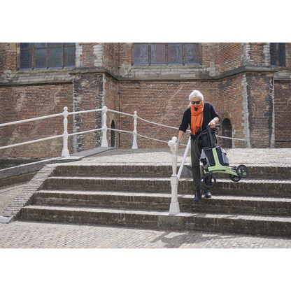 An older woman with white hair, sunglasses, and an orange scarf carefully descends stone steps outside a historic brick building, holding the handrail while using her Rollz Air Lightweight Folding Carbon Rollator, which weighs just 10.4 lbs.