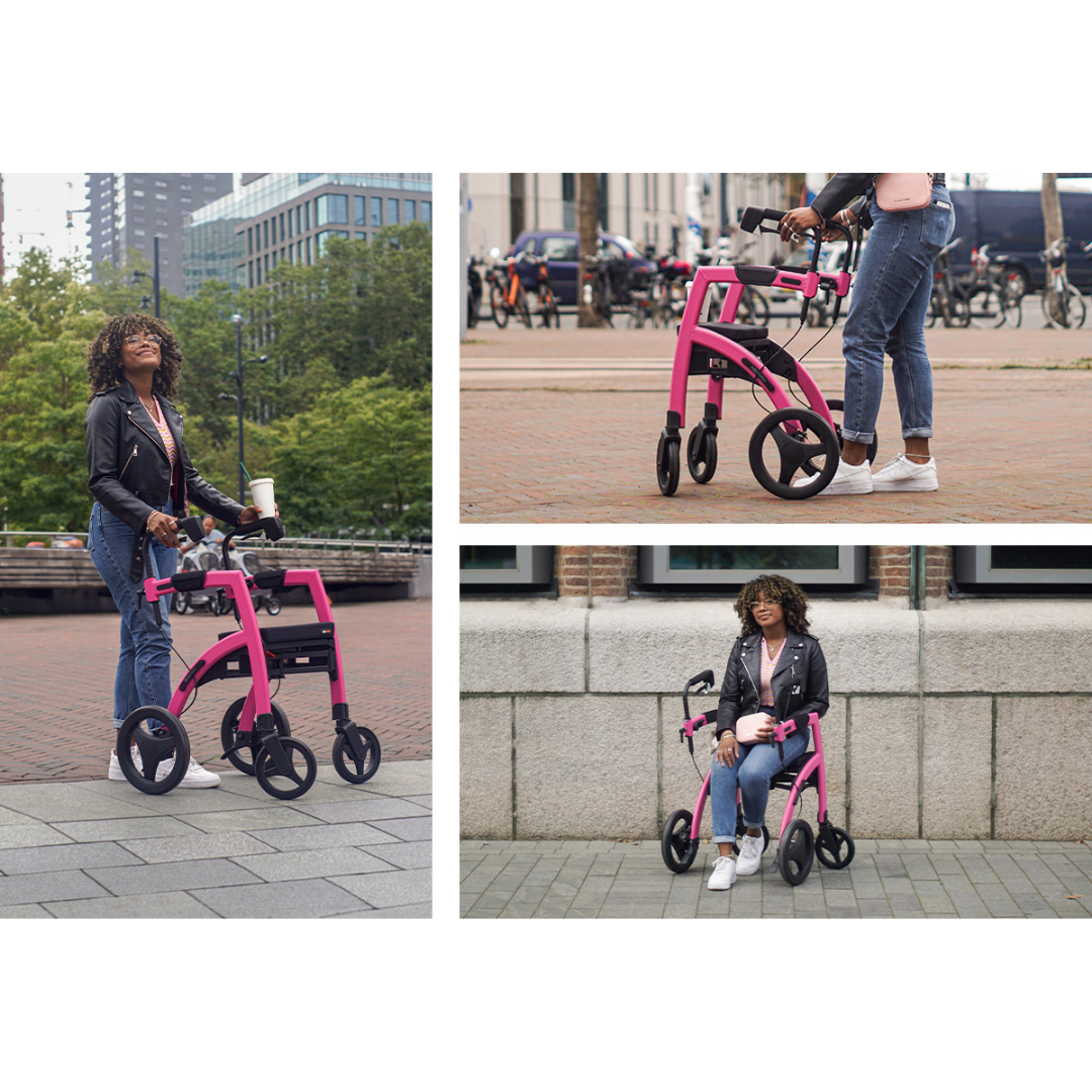 A woman with curly hair uses a bright pink Rollz Motion 2-in-1 Rollator Walker and Wheelchair outdoors in the city; she smiles, holding a coffee cup, and is casually dressed in jeans, a t-shirt, and a black jacket.