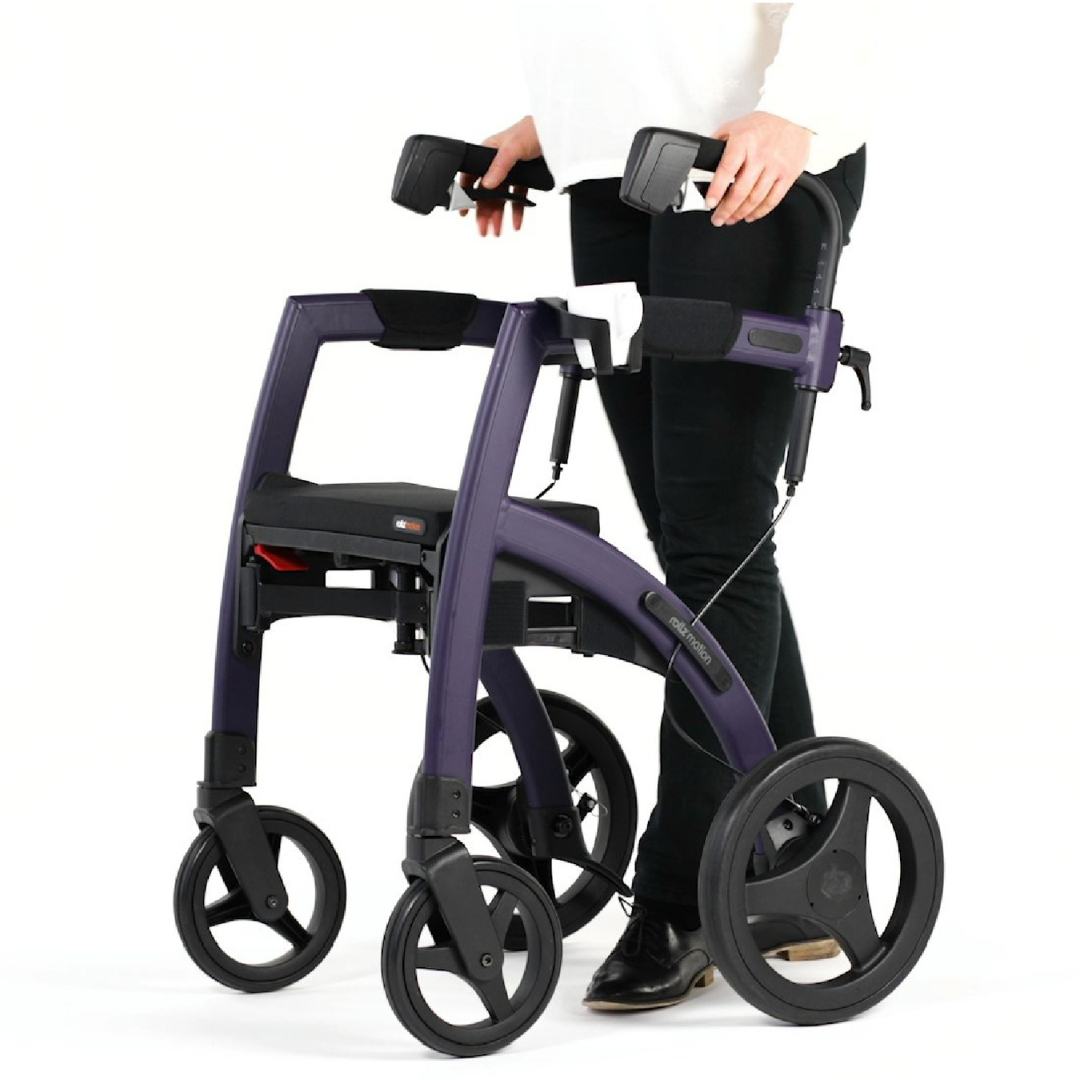 A person in black pants and a white shirt stands next to a sleek purple Rollz Motion Rollator, equipped with four wheels, hand brakes, and a versatile wheelchair package from the Rollz Motion Rollator Accessories for added convenience.