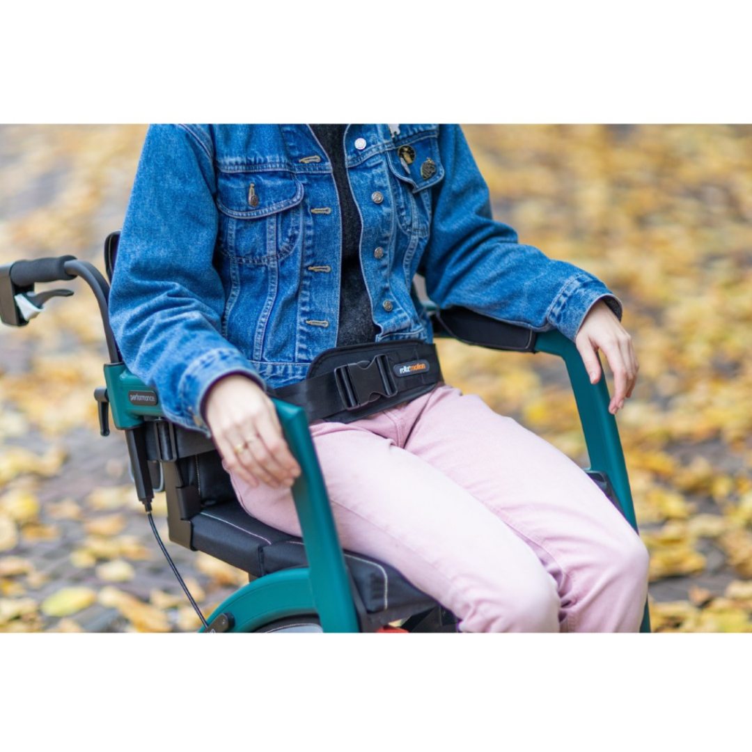 Seated in a wheelchair on a leafy path, someone wears a denim jacket and pink pants. The Rollz Motion Rollator, with teal armrests and a black seatbelt, perfectly complements the autumn atmosphere.