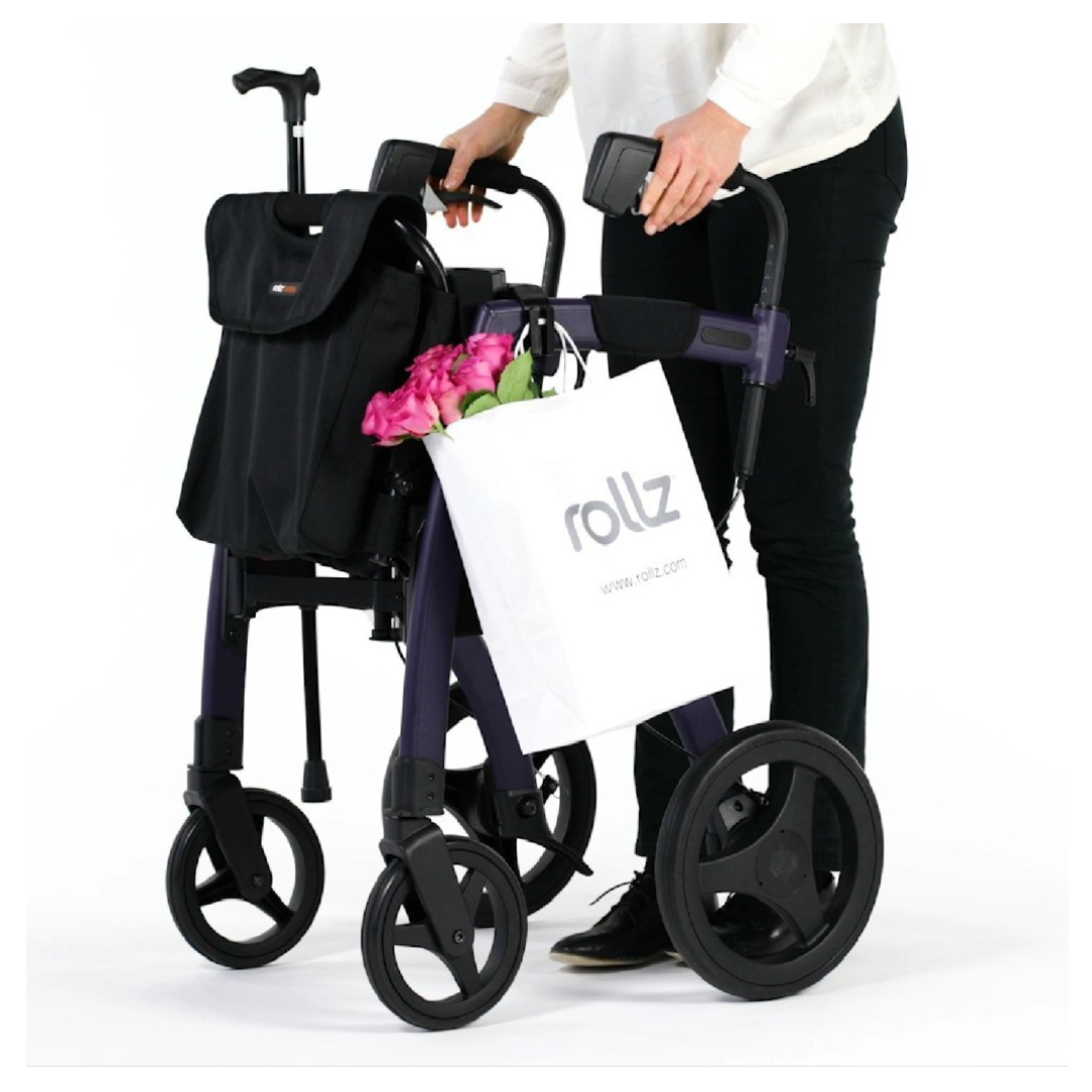 A person with a Rollz Motion Rollator, featuring a sleek foldable design and large wheels for smooth navigation, stands behind it holding pink roses and a white shopping bag. A black travel cover bag from the Rollz Motion Rollator Accessories is also attached.