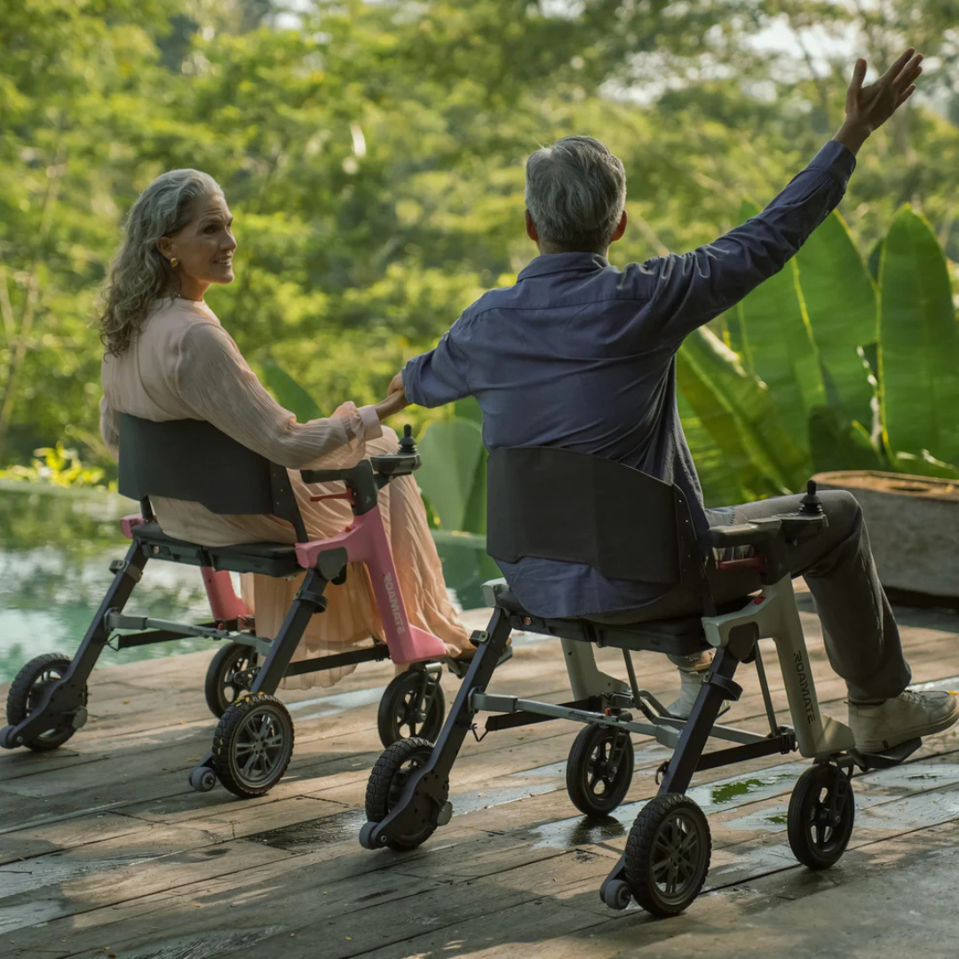 Two older adults relax outdoors on a wooden deck surrounded by greenery. The woman smiles at the man, who gestures energetically from his Roamate 3-in-1 Electric Rollator Wheelchair in the sunny, peaceful setting filled with connection and joy.