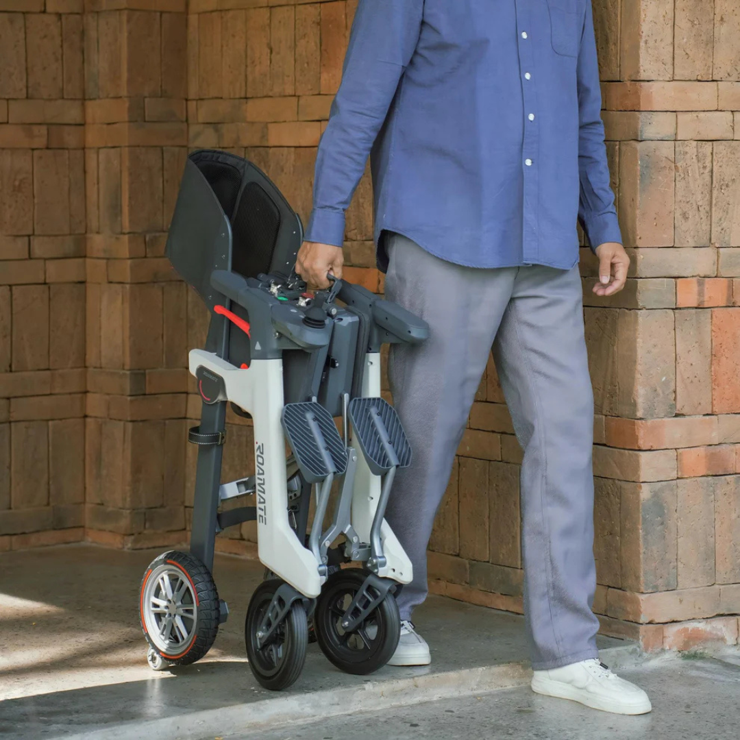 A person in a blue shirt and gray pants walks near a brick wall, carrying a folded Roamate 3-in-1 Electric Rollator Wheelchair by its frame, demonstrating convenient portability and versatile mobility.