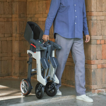 A person in a blue shirt and gray pants walks near a brick wall, carrying a folded Roamate 3-in-1 Electric Rollator Wheelchair by its frame, demonstrating convenient portability and versatile mobility.