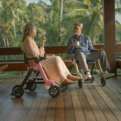 An older woman and man relax on a wooden deck in Roamate 3-in-1 Electric Rollator Wheelchairs, chatting and enjoying drinks amid lush tropical greenery and palm trees.