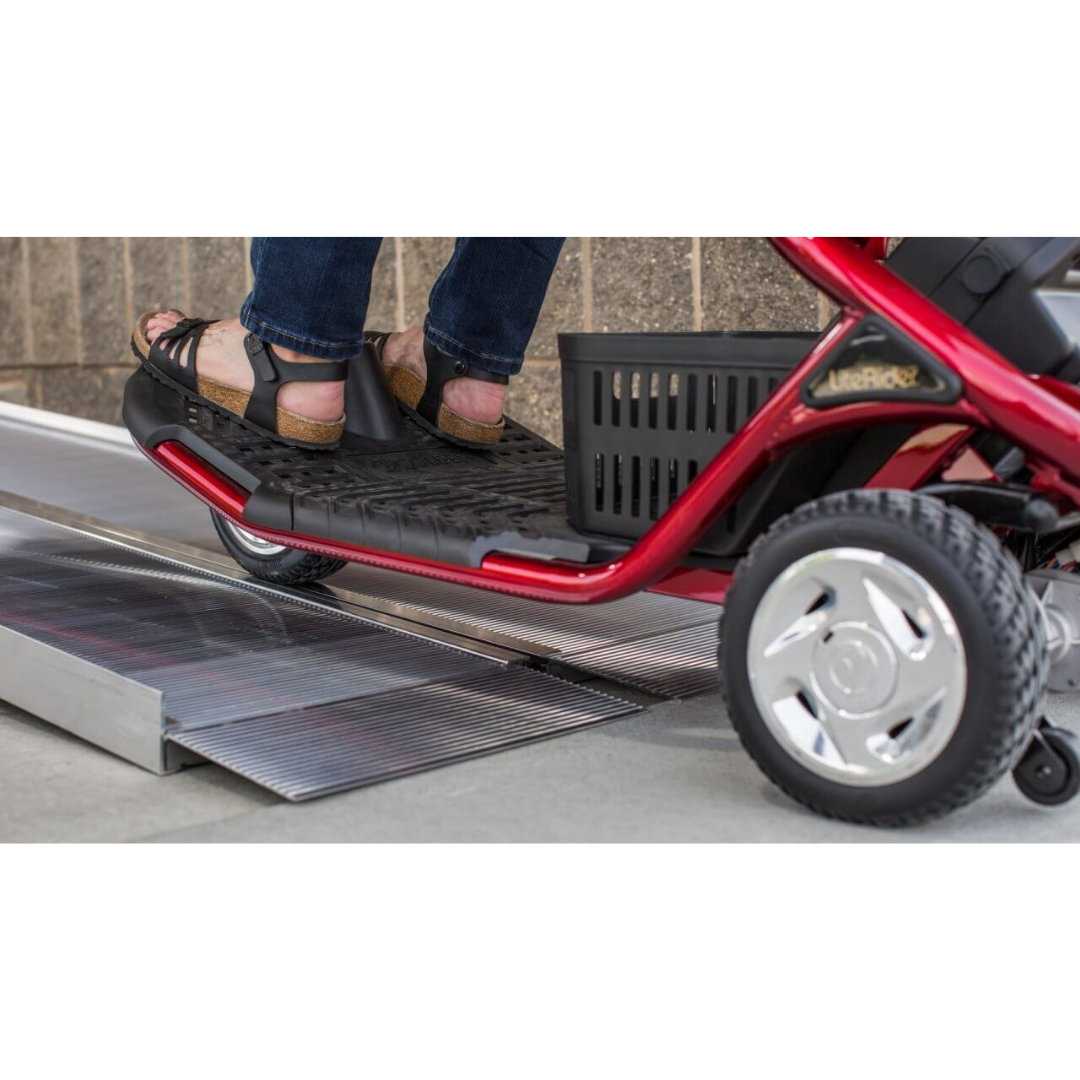 Close-up of a person’s feet on the footrest of a red mobility scooter going up the EZ-Access SUITCASE® Singlefold Mobility Ramp—portable metal ramp placed beside a brick wall. See videos or specs for details.