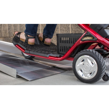 Close-up of a person’s feet on the footrest of a red mobility scooter going up the EZ-Access SUITCASE® Singlefold Mobility Ramp—portable metal ramp placed beside a brick wall. See videos or specs for details.