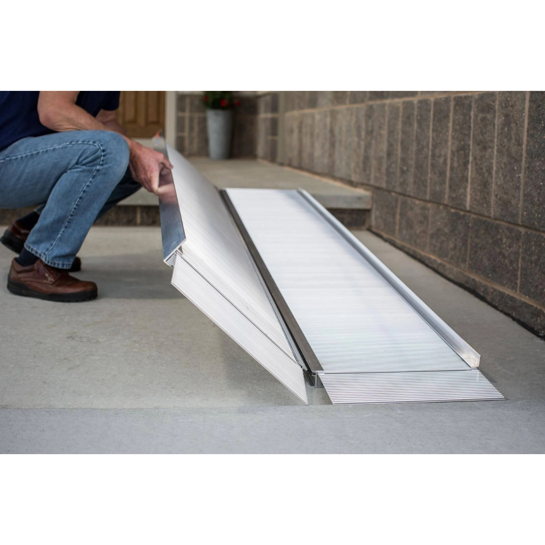 A person in jeans and brown shoes sets up the EZ-Access SUITCASE® Singlefold Mobility Ramp - Portable on a concrete step by a doorway, demonstrating its ease of use as shown in instructional videos.