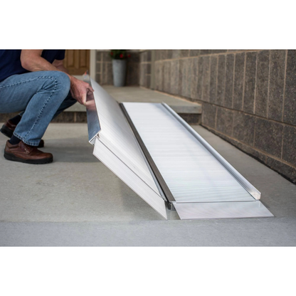 A person in jeans and brown shoes sets up the EZ-Access SUITCASE® Singlefold Mobility Ramp - Portable on a concrete step by a doorway, demonstrating its ease of use as shown in instructional videos.