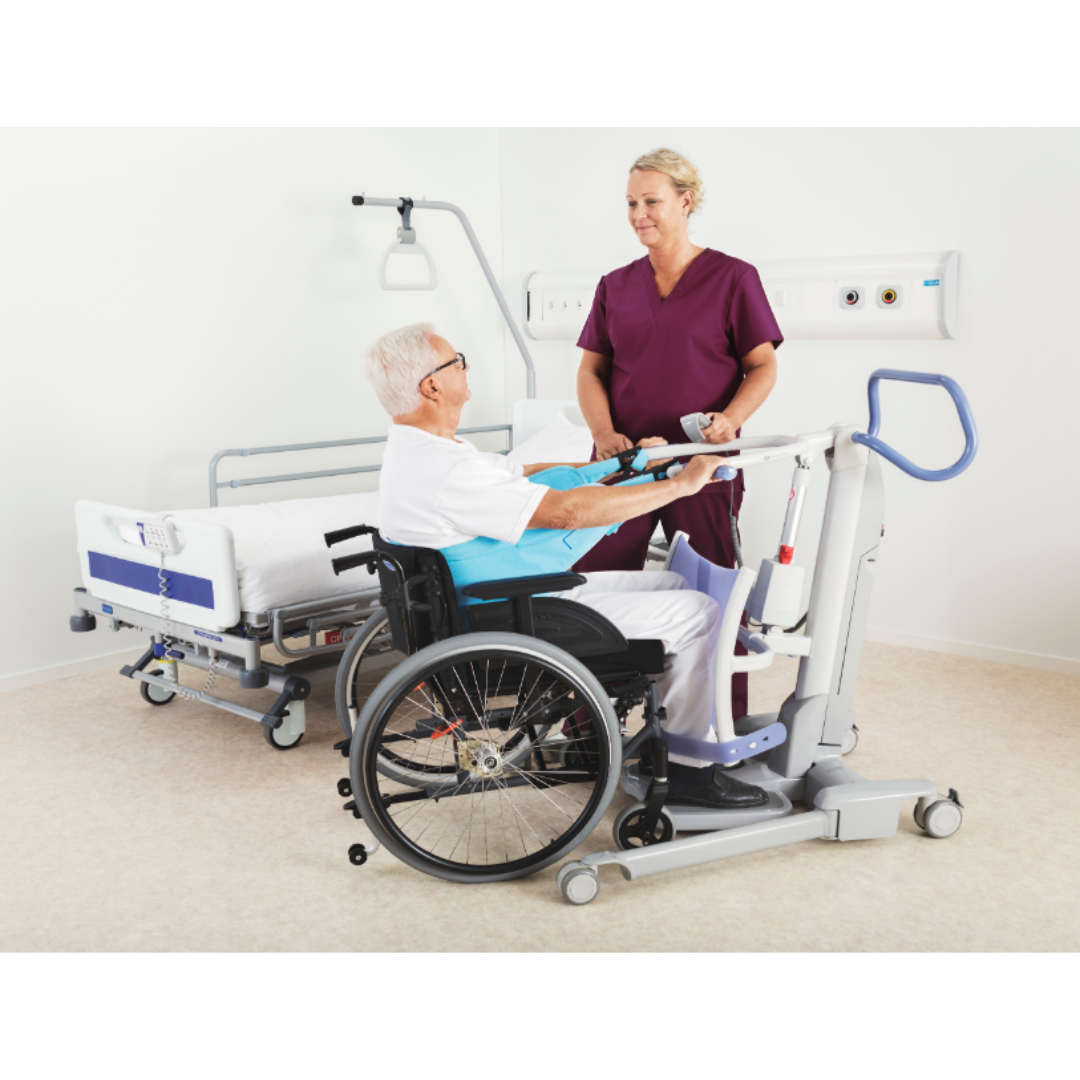 A healthcare worker uses the ARJO Sara Flex Sit-to-Stand Patient Transfer Lift to help an older man in a wheelchair transfer safely beside a hospital bed in a clinical setting.