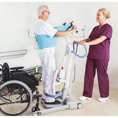 An elderly man in a support harness uses the ARJO Sara Flex Sit-to-Stand Patient Transfer Lift for a sit-to-stand transfer, assisted by a healthcare worker in purple scrubs. A wheelchair and hospital bed are nearby in this clinical setting.