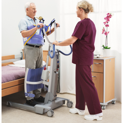An elderly man uses the ARJO Sara Flex Sit-to-Stand Patient Transfer Lift beside a bed, assisted by a healthcare worker in burgundy scrubs. A bedside cabinet with a potted orchid appears in the background.