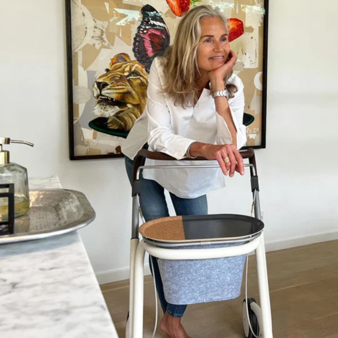 A smiling woman with long gray hair uses the sleek byACRE Scandinavian Butler At-Home Rollator in a bright room. Art of a lion and butterfly decorates the wall behind her, with a marble counter and soap in front.