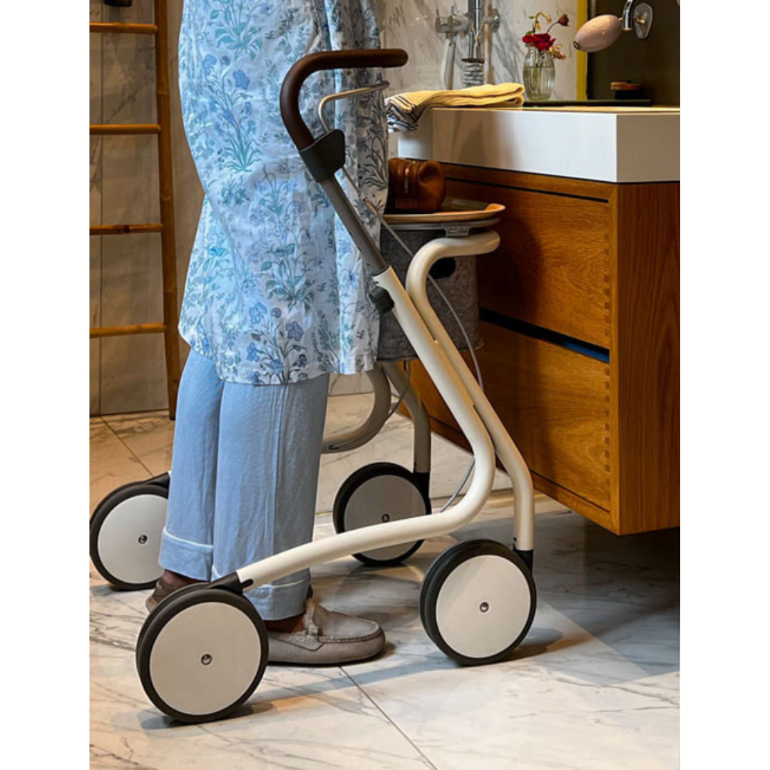An older adult in light blue pants and a floral tunic stands in a bathroom using the byACRE Scandinavian Butler At-Home Rollator, a modern white and black four-wheeled walker, near a wooden vanity.