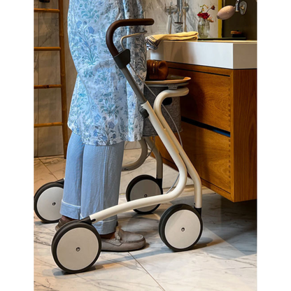 An older adult in light blue pants and a floral tunic stands in a bathroom using the byACRE Scandinavian Butler At-Home Rollator, a modern white and black four-wheeled walker, near a wooden vanity.