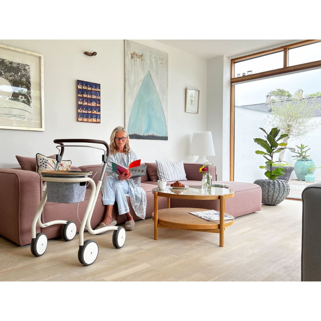 An older woman sits on a pink sofa reading a red book in a bright, modern living room. Her byACRE Scandinavian Butler At-Home Rollator rests beside her, matching the coffee table with flowers, artwork, and large sunlit windows.