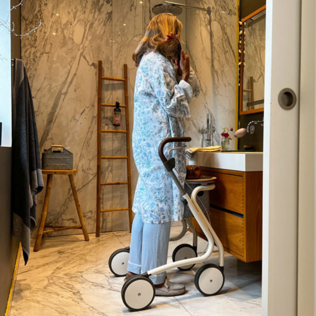 A woman in pajamas stands in a bathroom with marble walls and modern fixtures, using her byACRE Scandinavian Butler At-Home Rollator while holding a small dog and looking into the mirror.