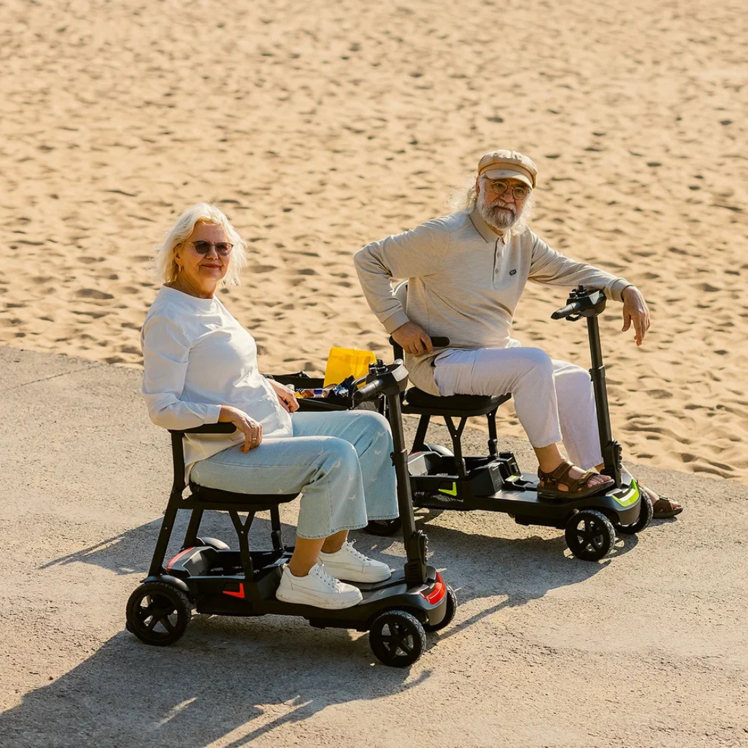 Two older adults ride ScootNGo Lite Portable Folding Scooters on a paved path by the beach, smiling in light clothing and sunglasses—highlighting the scooters’ comfort and high-quality design.