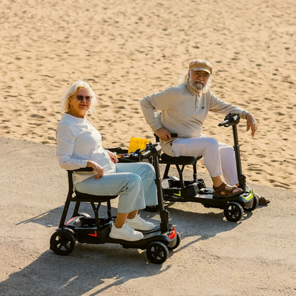 Two older adults ride ScootNGo Lite Portable Folding Scooters on a paved path by the beach, smiling in light clothing and sunglasses—highlighting the scooters’ comfort and high-quality design.