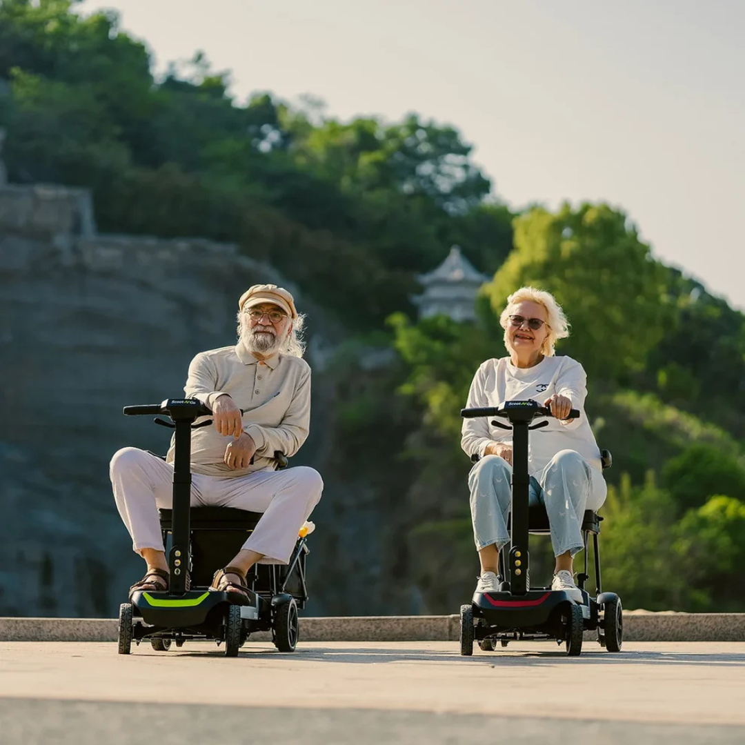 Two older adults, smiling and relaxed, sit outdoors on ScootNGo Lite Portable Folding Scooters. Enjoying the scenic backdrop of trees and hills, they showcase how this reliable scooter is perfect for leisurely outings.