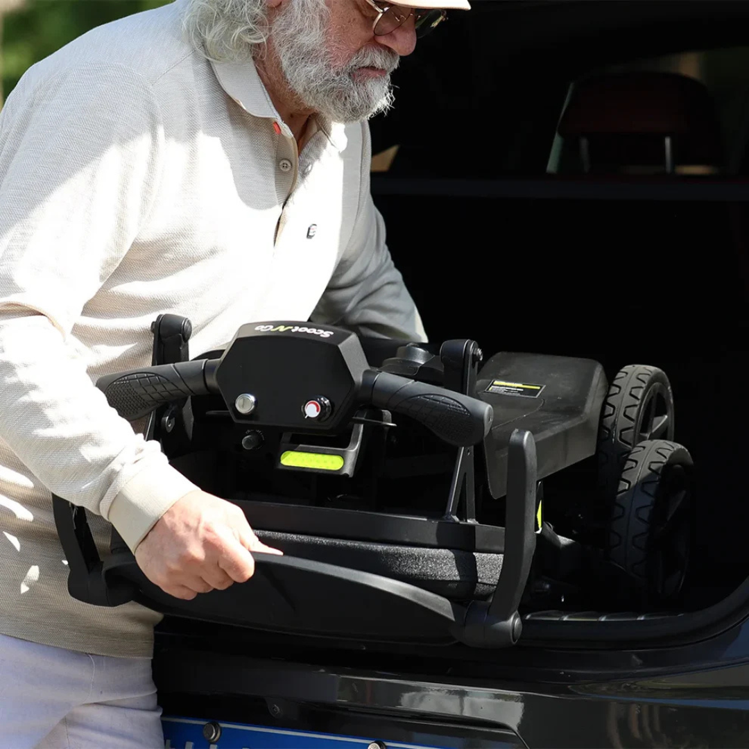 An older man with gray hair and a beard, wearing sunglasses and a beige long-sleeve shirt, places the ScootNGo Lite Portable Folding Scooter—renowned for its impressive specs—into his car trunk.
