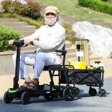 An elderly man with white hair and beard, in a beige cap and sunglasses, rides a ScootNGo Lite Portable Folding Scooter, towing a wagon of groceries along a sunny outdoor path.