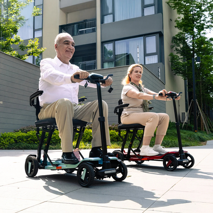 An older man and woman ride ScootNgo Voyager Lightweight Folding Scooters outdoors on a sunny day, smiling as they travel side by side on a paved path in front of a modern apartment building.