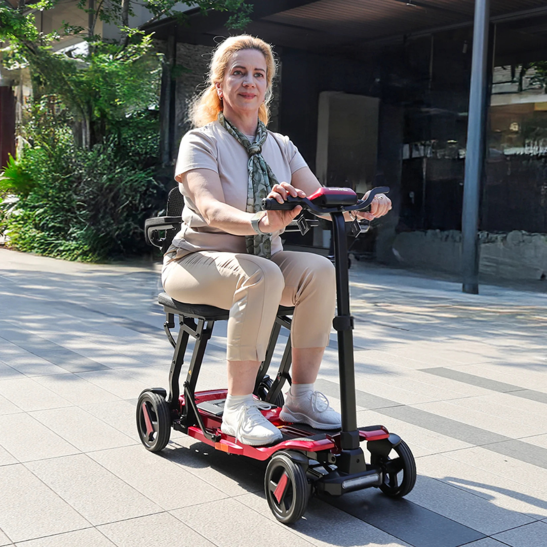 A light-haired woman in a pale outfit and scarf confidently rides a ScootNgo Voyager Lightweight Folding Scooter along a sunny sidewalk, with greenery and modern buildings visible in the background.