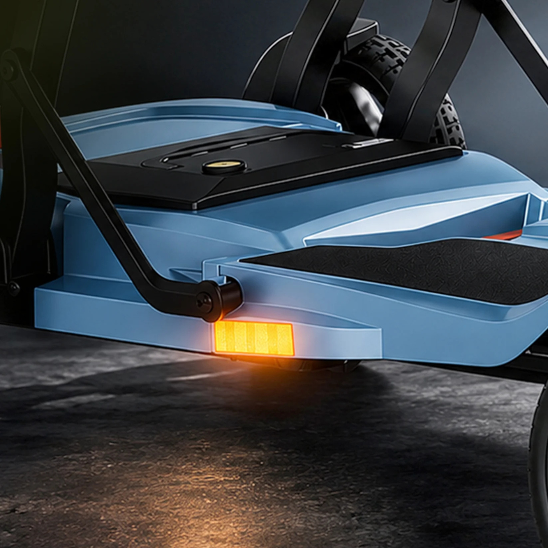 Close-up of the blue ScootNgo Voyager Lightweight Folding Scooter’s footboard and rear wheel, showing an illuminated orange side reflector and part of the suspension system, displayed on a dark surface.