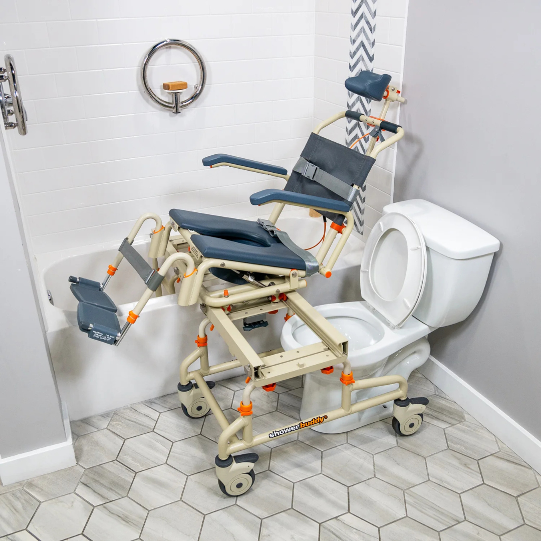 The ShowerBuddy SB2T TubBuddy with Tilt, in beige and gray, is shown positioned over a white toilet with part extending into a bathtub. Tiled flooring and safety bars highlight its accessible design for safe and convenient bathing.