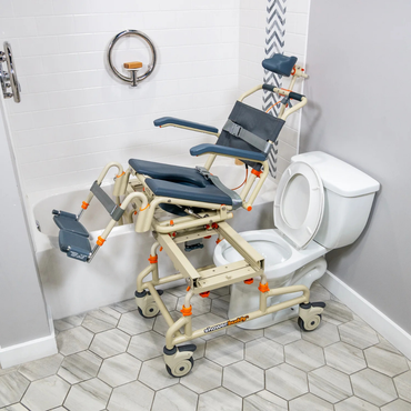 The ShowerBuddy SB2T TubBuddy with Tilt, in beige and gray, is shown positioned over a white toilet with part extending into a bathtub. Tiled flooring and safety bars highlight its accessible design for safe and convenient bathing.