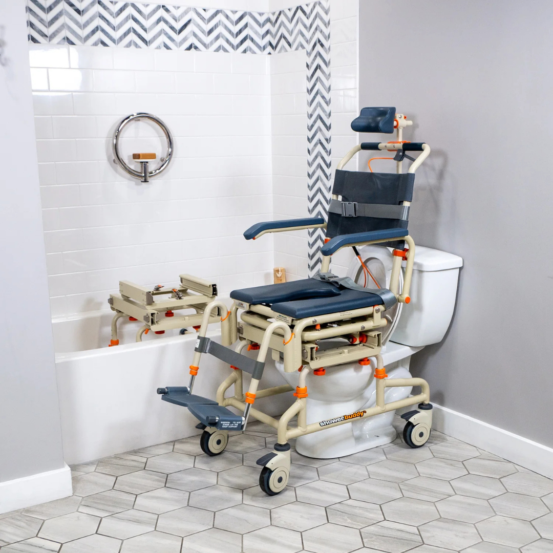 The ShowerBuddy SB2T TubBuddy with Tilt is positioned over a toilet beside a bathtub with a grab bar and bath transfer bench in a tiled bathroom, creating an accessible bathing system.
