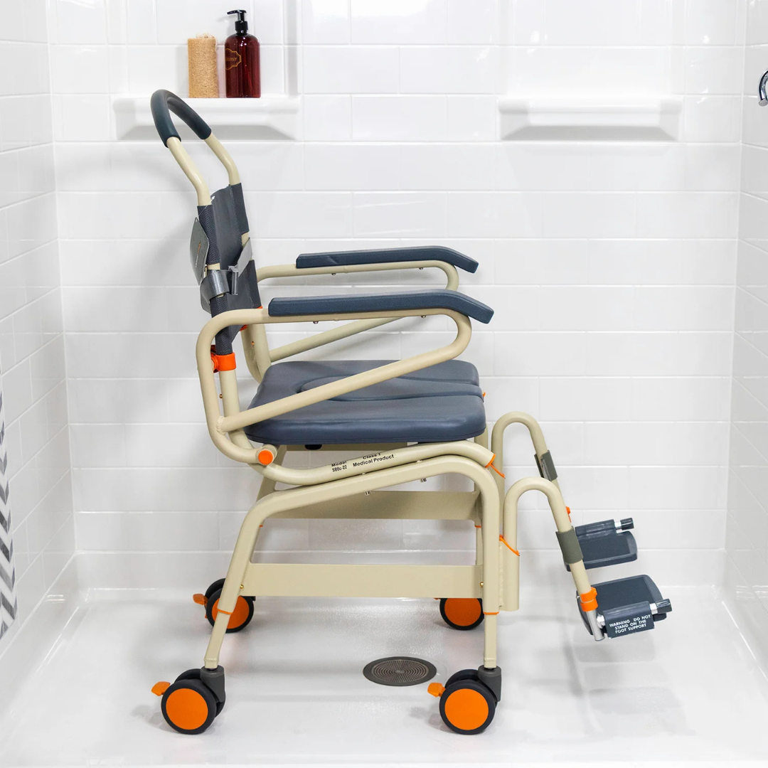 The ShowerBuddy SB6C Bariatric Rolling Shower Chair, featuring a padded seat, armrests, footrests, orange accents, and wheels for mobility, sits in a white-tiled shower stall to provide comfort and support while bathing.