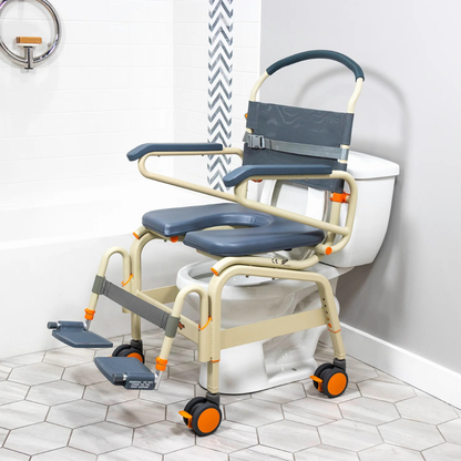 The ShowerBuddy SB6C Bariatric Rolling Shower Chair, with padding, armrests, footrests, and wheels, is shown over a toilet in a modern bathroom featuring hexagonal floor tiles and a white bathtub.