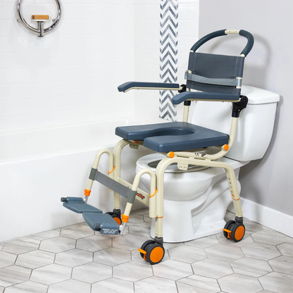 The ShowerBuddy SB6C Roll-in shower chair Commode, featuring gray and beige colors with orange accents, is shown over a toilet in a white-tiled bathroom. It includes wheels, armrests, and a footrest for accessibility and mobility.